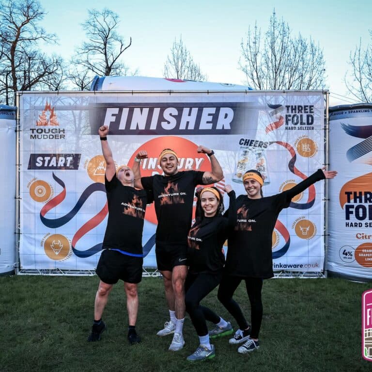 Four team members celebrating in front of the finisher banner at a Tough Mudder event.