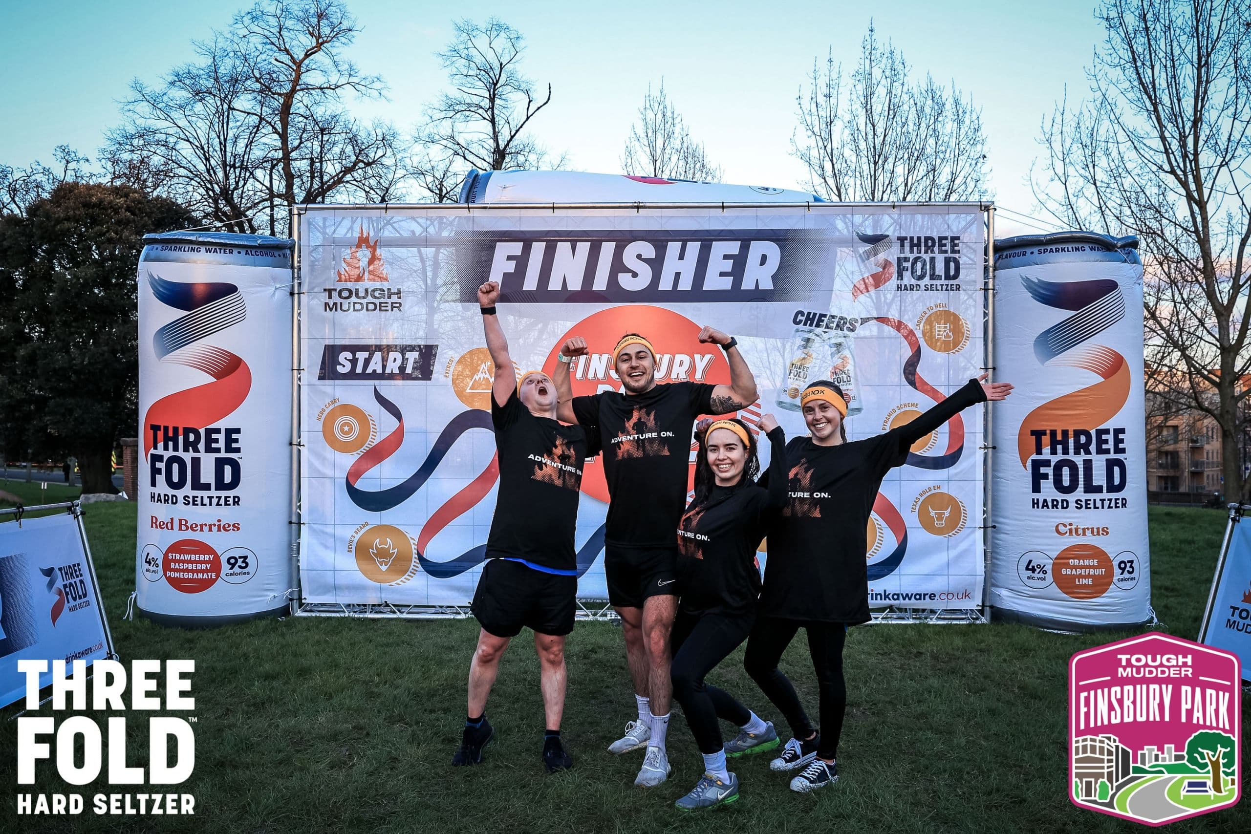 Tough Mudder 5k Four team members celebrating in front of the finisher banner at a Tough Mudder event, wearing matching black event t-shirts and orange headbands, flexing and cheering with arms raised.