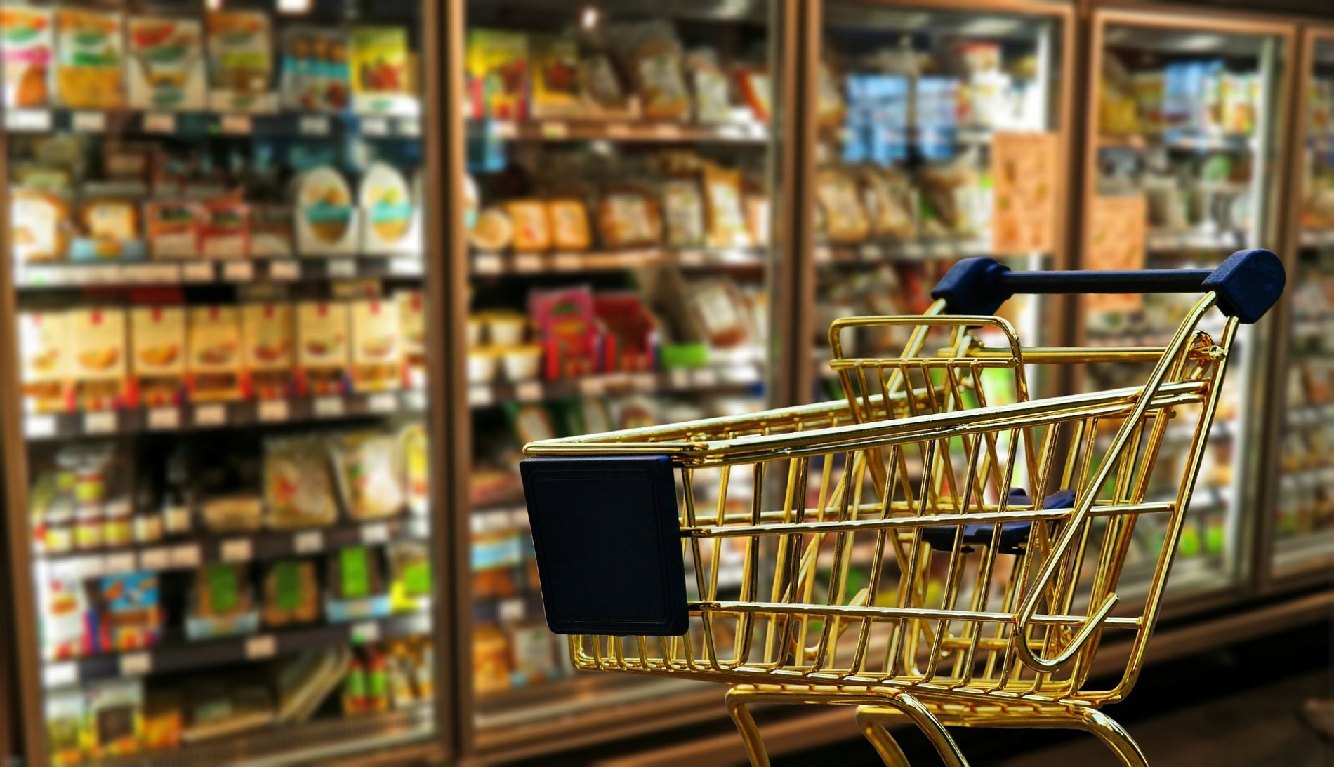 A miniature shopping trolley sits in front of fridges and freezers in a super market aisle