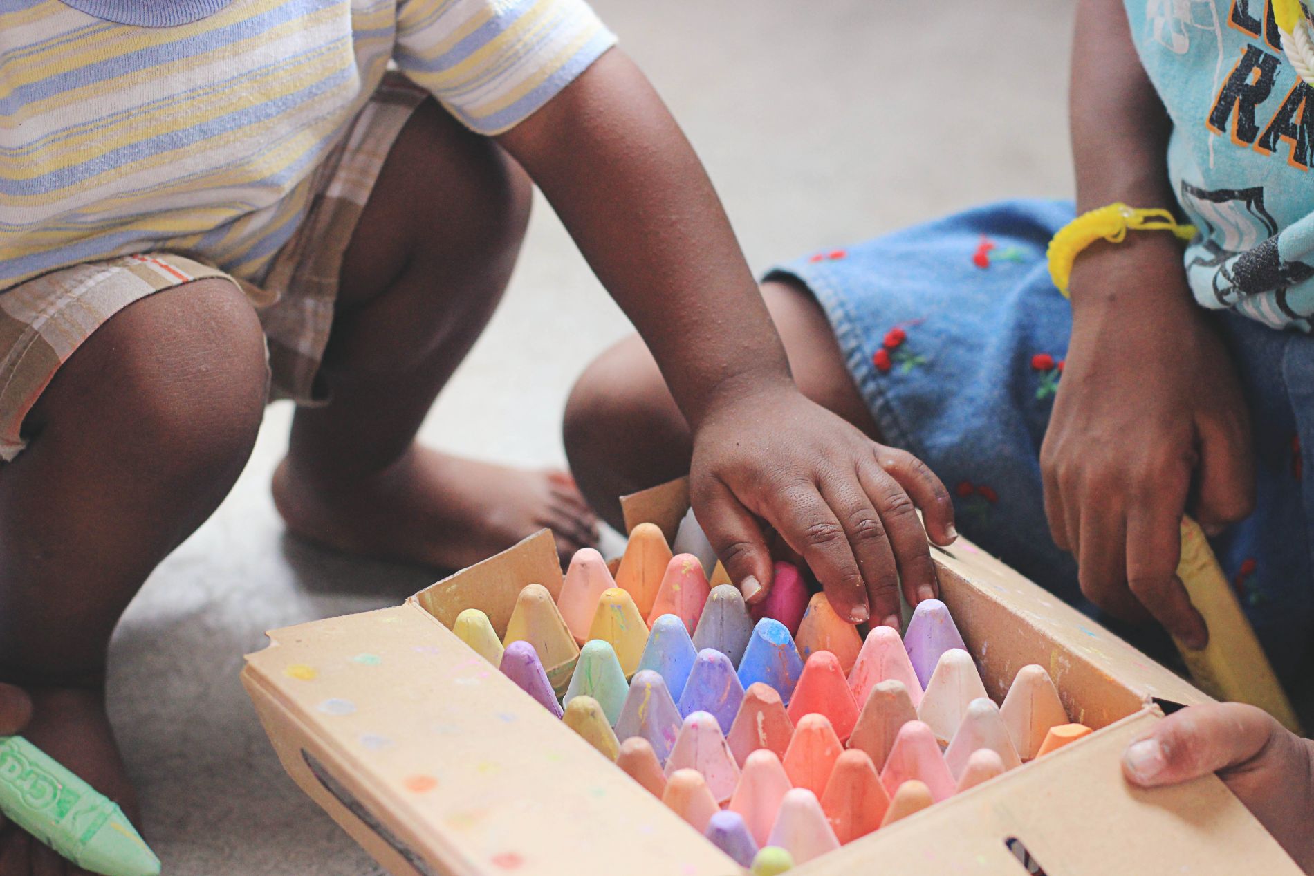 A young child looks through a box of colourful chalks next to another child holding the box