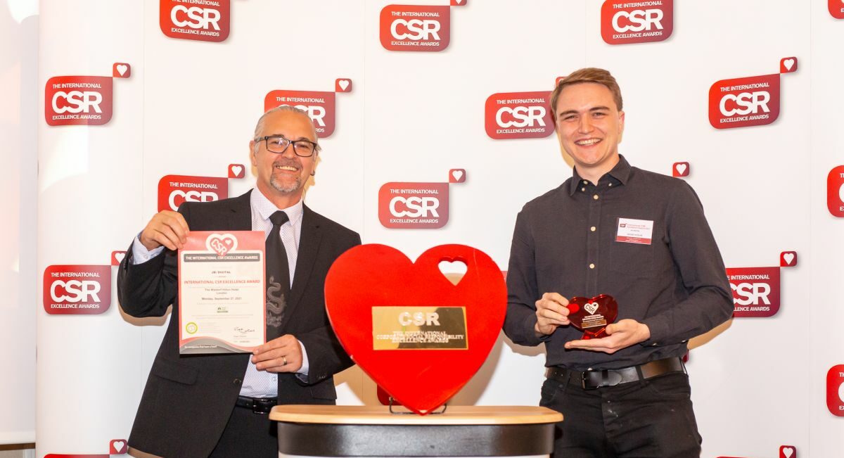 Two JBi team members smiling at the International CSR Excellence Awards, one holding the award certificate and the other a heart-shaped trophy, in front of the event's branded backdrop.