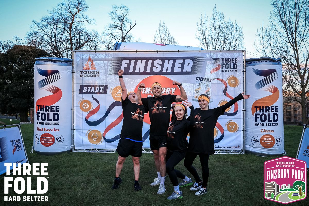 Four team members celebrating in front of the finisher banner at a Tough Mudder event, wearing matching black event t-shirts and orange headbands, flexing and cheering with arms raised.