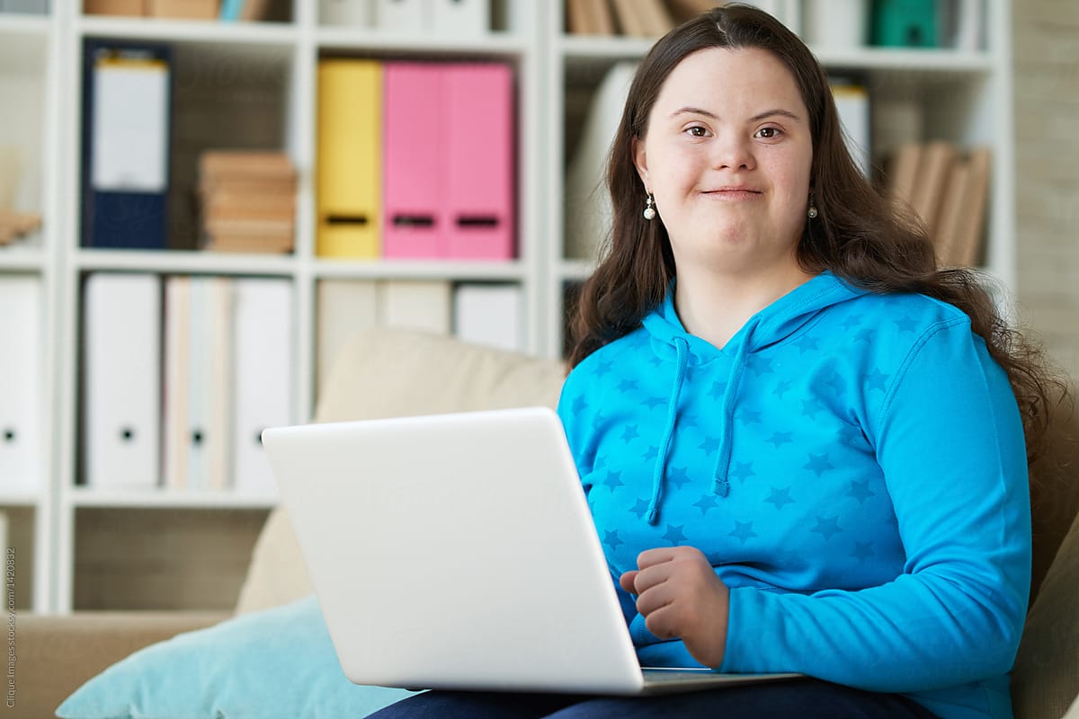 Young woman with Down syndrome smiling while using a laptop. She is wearing a bright blue hoodie with star patterns and sitting in a comfortable room with shelves full of colourful binders and books in the background.